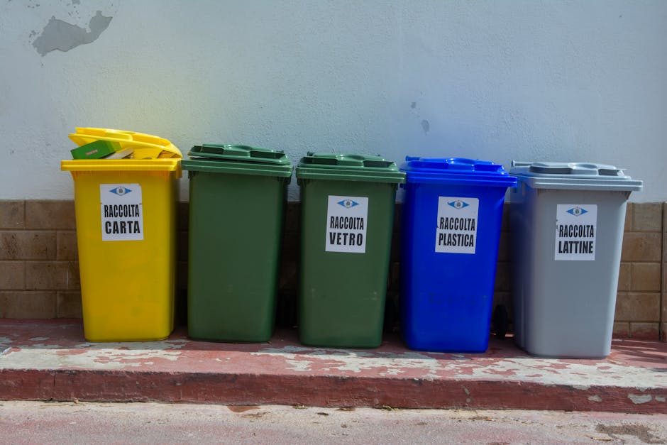 A lineup of five colour-coded recycling bins positioned on a paved sidewalk against a white wall with a brick base. The first bin on the left is yellow with a slightly open lid, displaying a collection of yellow plastic items inside; it has a label reading 'RACCOLTA CARTA' indicating paper recycling. To its right is a tall, dark green bin with a closed lid, marked 'RACCOLTA VETRO' for glass collection. Next is another similar green bin, also with a closed lid, designated for glass as well. The fourth bin is blue, with a closed lid and labeled 'RACCOLTA PLASTICA' for plastic recycling. The last bin on the right is light grey with a closed lid, marked 'RACCOLTA LATTINE' for tin cans. The scene appears to be in an outdoor urban or residential area, likely related to private waste collection services, with the bins situated on a slightly elevated curb. The lighting is natural, casting soft shadows on the ground and emphasizing the different textures of the smooth plastic surfaces of the bins and the wall background. This arrangement exemplifies organised waste segregation using dedicated refuse containers, supporting efficient rubbish removal and recycling efforts in the local area.