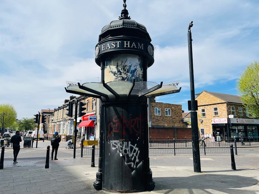 A cylindrical glass and metal information kiosk with a domed, decorative black metal top situated at a busy pedestrian intersection on Clapham High Street. The kiosk features a clock face, with the words 'EAST HAM' visible on a curved sign just below the dome, indicating a historical or directional marker. The lower section of the kiosk is made of black metal mesh surrounded by graffiti tags in red and white spray paint. The surrounding environment includes black metal bollards lining the pavement, with a few people walking nearby, some dressed in dark clothing. In the background, there are small retail shops with signage, including a nail salon and a beauty store, along with residential buildings constructed from brick and stone. To the right, a tall black lamppost stands beside the kiosk, and a bright, partly cloudy sky illuminates the scene, creating a clear daytime atmosphere typical of urban streets. This scene highlights the mix of historic street furniture and modern urban surroundings commonly associated with alternative waste handling and civil infrastructure in the area, as managed by local services such as Rubbish Collection Clapham.