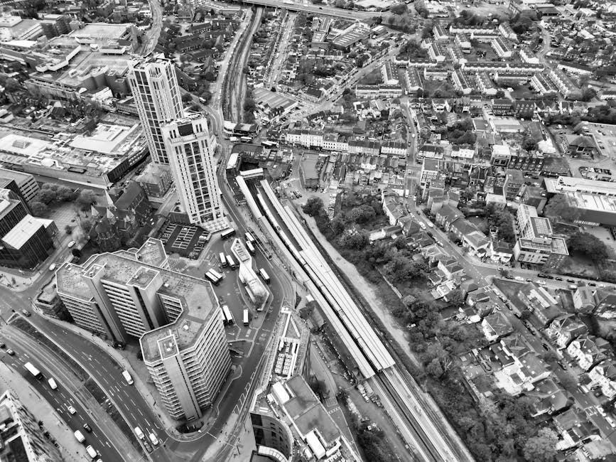 An aerial black-and-white photograph shows a dense urban area with a mixture of modern high-rise buildings and older, smaller residential houses. In the foreground, several multi-storey office buildings and apartments with flat roofs and glass windows are arranged along curved roads filled with moving vehicles. A railway line runs diagonally across the image, with a long, elevated train track and a stopped train visible. Surrounding the railway, there are many tightly packed houses with pitched roofs, narrow streets, and small gardens, indicating a residential neighbourhood. The background features additional streets, buildings, and green areas, with some trees and greenery scattered throughout the urban landscape. The overall scene highlights a busy city centre with mixed commercial, residential, and transport infrastructure, typical of a densely populated metropolitan environment. This depiction supports the context of independent waste collection and on-site clearance services by illustrating a built-up area where proper rubbish removal and management are essential, with Rubbish Collection Clapham operating within such environments.