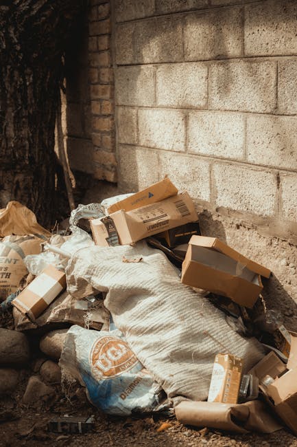 A pile of mixed waste materials accumulated at the base of a garden wall, including flattened cardboard boxes, plastic bags, and crumpled paper packaging. The cardboard boxes vary in size and are partially opened, revealing their contents. The plastic bags are light brown and beige, appearing loosely filled, with some material spilling out. The surrounding environment features a rough, grey concrete brick wall in the background, with a small section of darker, weathered brickwork visible in the upper left corner, suggesting an outdoor or semi-enclosed area. The ground surface is composed of dirt and small stones, with shadows cast from nearby objects, indicating natural daylight. The scene exemplifies typical waste that may require professional rubbish removal, such as that provided by companies like Rubbish Collection Clapham, especially for private property clearance or alternative disposal options outside local authority services.