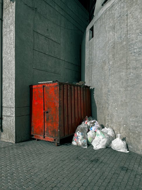 A red metal skip with a weathered, rusted surface positioned against a dark teal-colored wall and a light gray concrete wall on a paved outdoor surface. The skip contains partially visible waste, and several white plastic bags filled with rubbish are piled next to it, some leaning against the skip's lower edge. The scene appears to be on a narrow alleyway or side street, with minimal natural light. The concrete wall has a textured surface, and the dark teal wall features horizontal paneling, providing a contrasting background. Visible in the background is a small rectangular window opening high on the teal wall, with no glass or covering. The overall scene depicts a typical private waste collection area, aligned with the types of rubbish disposal handled by waste management services such as Rubbish Collection Clapham, illustrating on-site clearance or alternative waste handling options.