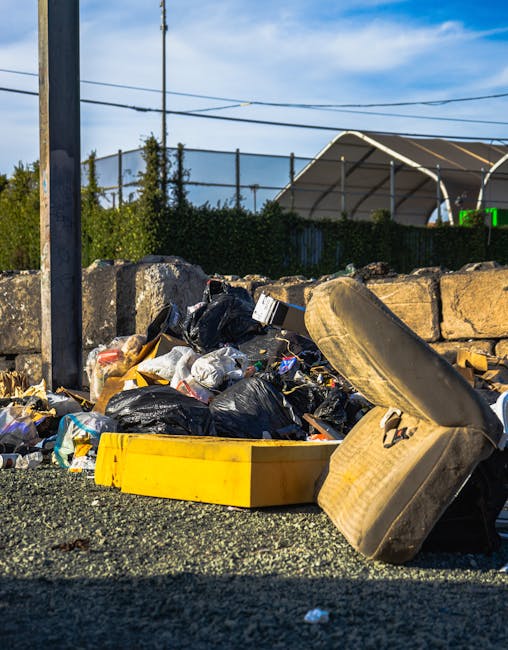 A pile of mixed waste materials accumulated at the base of a garden wall, including flattened cardboard boxes, plastic bags, and crumpled paper packaging. The cardboard boxes vary in size and are partially opened, revealing their contents. The plastic bags are light brown and beige, appearing loosely filled, with some material spilling out. The surrounding environment features a rough, grey concrete brick wall in the background, with a small section of darker, weathered brickwork visible in the upper left corner, suggesting an outdoor or semi-enclosed area. The ground surface is composed of dirt and small stones, with shadows cast from nearby objects, indicating natural daylight. The scene exemplifies typical waste that may require professional rubbish removal, such as that provided by companies like Rubbish Collection Clapham, especially for private property clearance or alternative disposal options outside local authority services.