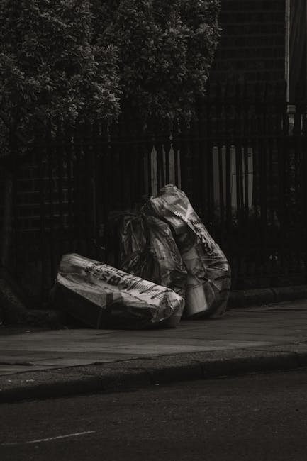 A pair of large black plastic rubbish bags filled with waste are placed on the pavement in front of a tall, black iron fence with vertical bars. The bags appear crumpled and slightly torn, with some loose material visible. Behind the fence, dense foliage from a leafy tree is visible, providing a natural green background. The scene is outdoors on a street with a concrete curb separating the pavement from the road, which is partially visible in the foreground. The lighting is subdued, suggesting an overcast day or low light conditions, and the overall scene reflects a typical outdoor rubbish disposal area that could be handled through independent collection or private waste removal services, such as those offered by Rubbish Collection Clapham, in line with local rubbish management practices.