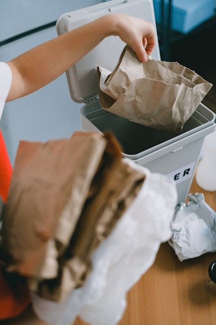 A person is seen disposing of brown paper bags into a white rubbish bin with a black lid, which is placed on a wooden surface indoors. The person's arm is extended over the bin, holding a paper bag open, ready to drop it inside. Several crumpled paper bags are in the foreground, while additional paper waste is visible nearby on the table. The environment appears to be a home or small office setting, with natural lighting illuminating the scene. The arrangement suggests an organised approach to waste disposal, potentially related to alternative rubbish collection methods or private waste management services provided by companies like Rubbish Collection Clapham, aiming to streamline household or small-scale rubbish removal processes visually depicted here.