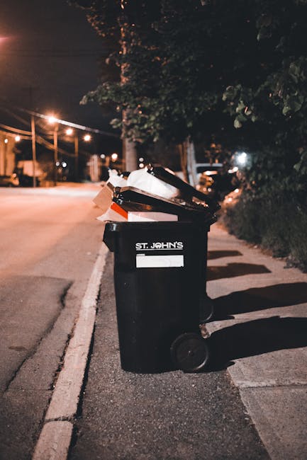A pair of large black plastic rubbish bags filled with waste are placed on the pavement in front of a tall, black iron fence with vertical bars. The bags appear crumpled and slightly torn, with some loose material visible. Behind the fence, dense foliage from a leafy tree is visible, providing a natural green background. The scene is outdoors on a street with a concrete curb separating the pavement from the road, which is partially visible in the foreground. The lighting is subdued, suggesting an overcast day or low light conditions, and the overall scene reflects a typical outdoor rubbish disposal area that could be handled through independent collection or private waste removal services, such as those offered by Rubbish Collection Clapham, in line with local rubbish management practices.